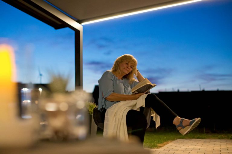 Frau liest entspannt ein Buch in der Abenddämmerung unter einer Pergola Markise mit LED Beleuchtung